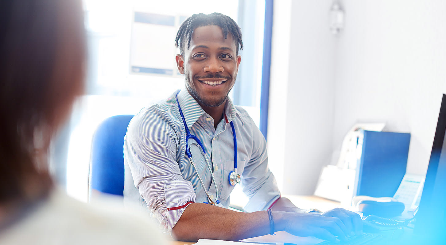 A healthcare provider at a desk with a laptop smiles as he interacts with a patient.